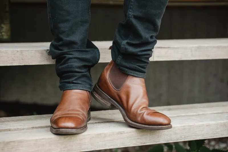 R.M. Williams Comfort Craftsman i brunt, til mørke jeans, hælen løftet midt i et steg på en tretrapp