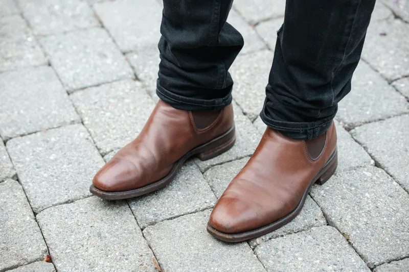 Brune R.M. Williams Comfort Craftsman til sorte jeans, sett ovenfra på grå brostein
