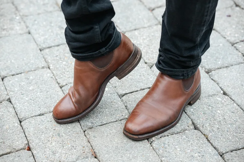 Brune R.M. Williams Comfort Craftsman til sorte jeans, tærne fremover på brostein