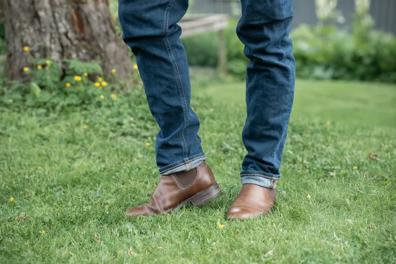 Brune R.M. Williams Comfort Craftsman til blå jeans, midt i et steg på gress