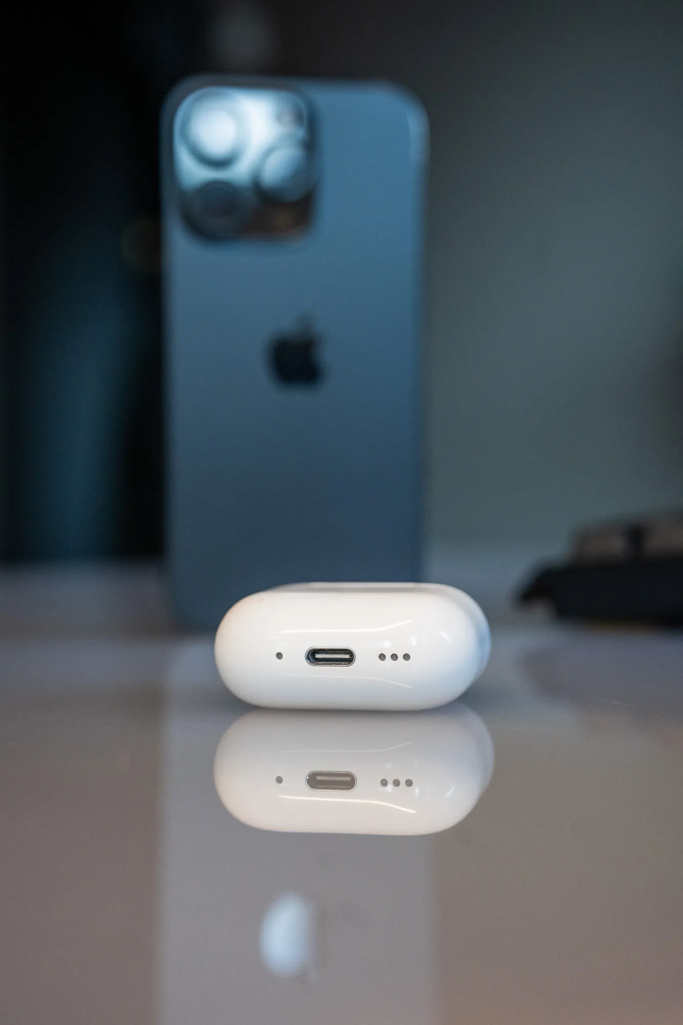 Undersiden av AirPods 5-etuiet med USB-C-port og høyttalerhull, en iPhone uskarp i bakgrunnen