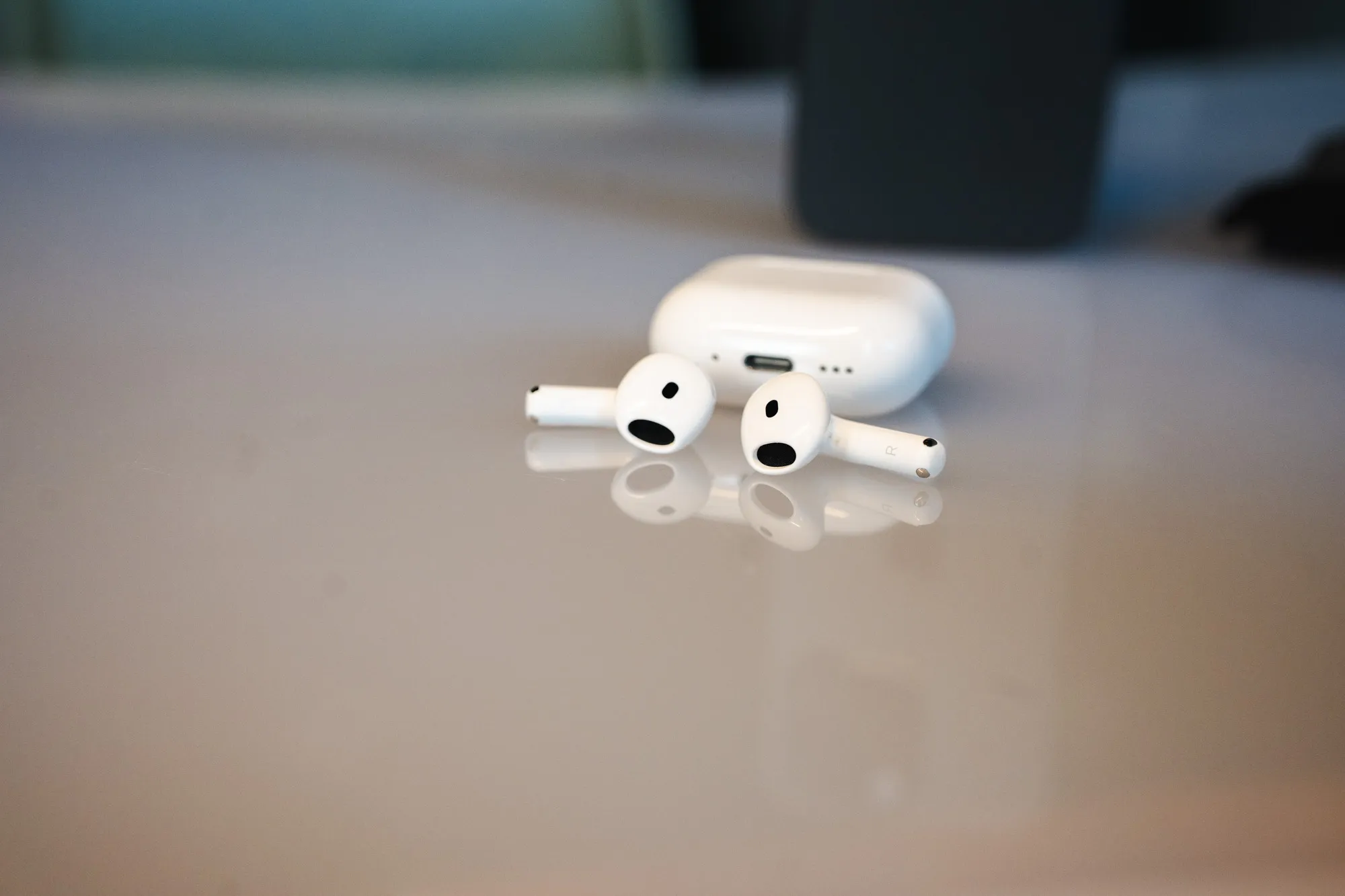 Begge AirPods 5-proppene ligger foran etuiet på et blankt bord, med USB-C-porten synlig på etuiet