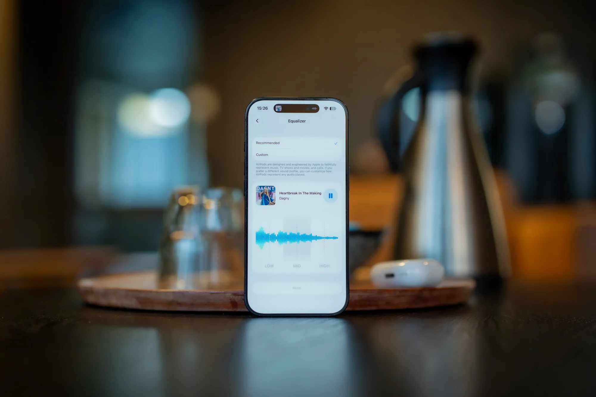 iPhone stående på et trebrett med equalizeren for AirPods 5 på skjermen, AirPods-etuiet og en termokanne i bakgrunnen
