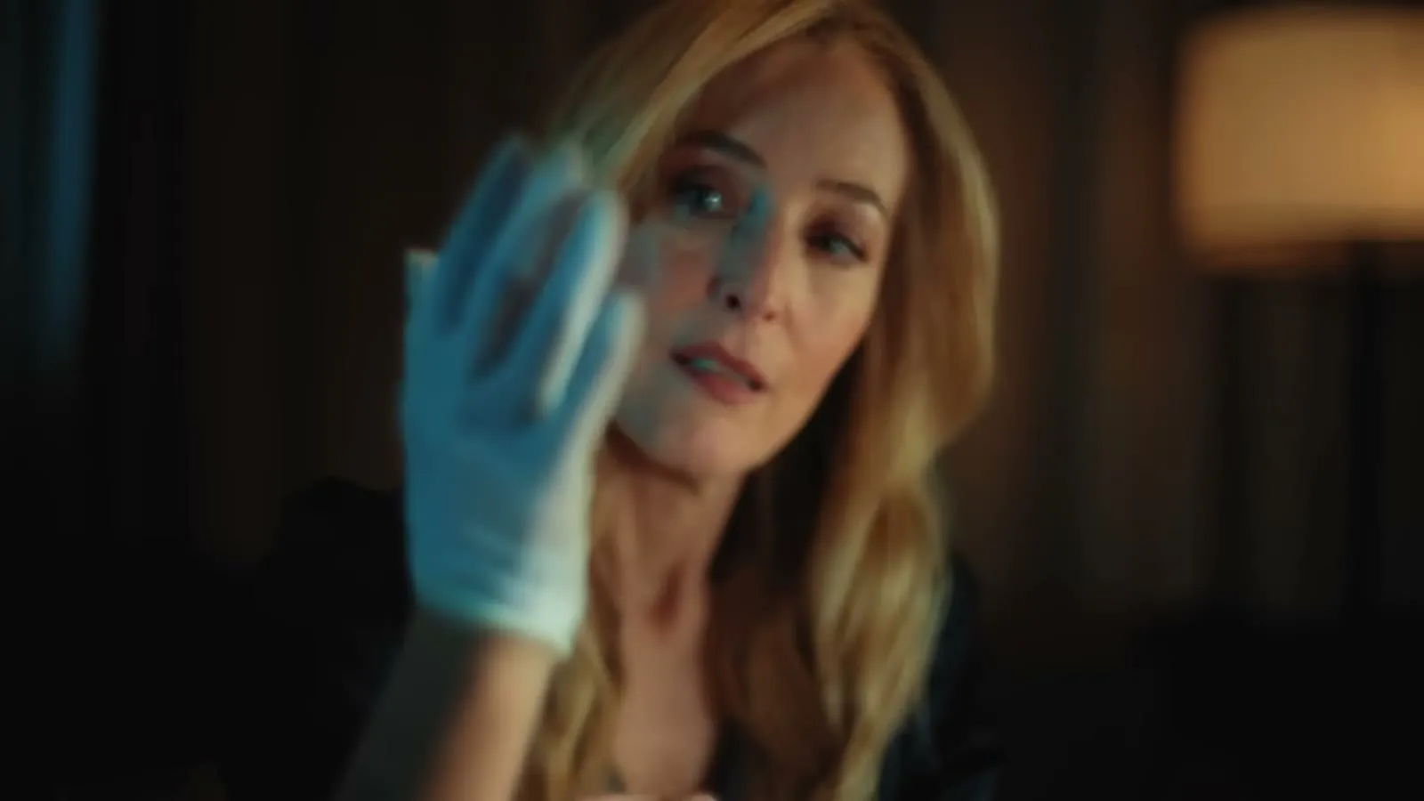 Gillian Anderson trekker på seg en blå latekshanske i et mørkt hotellrom